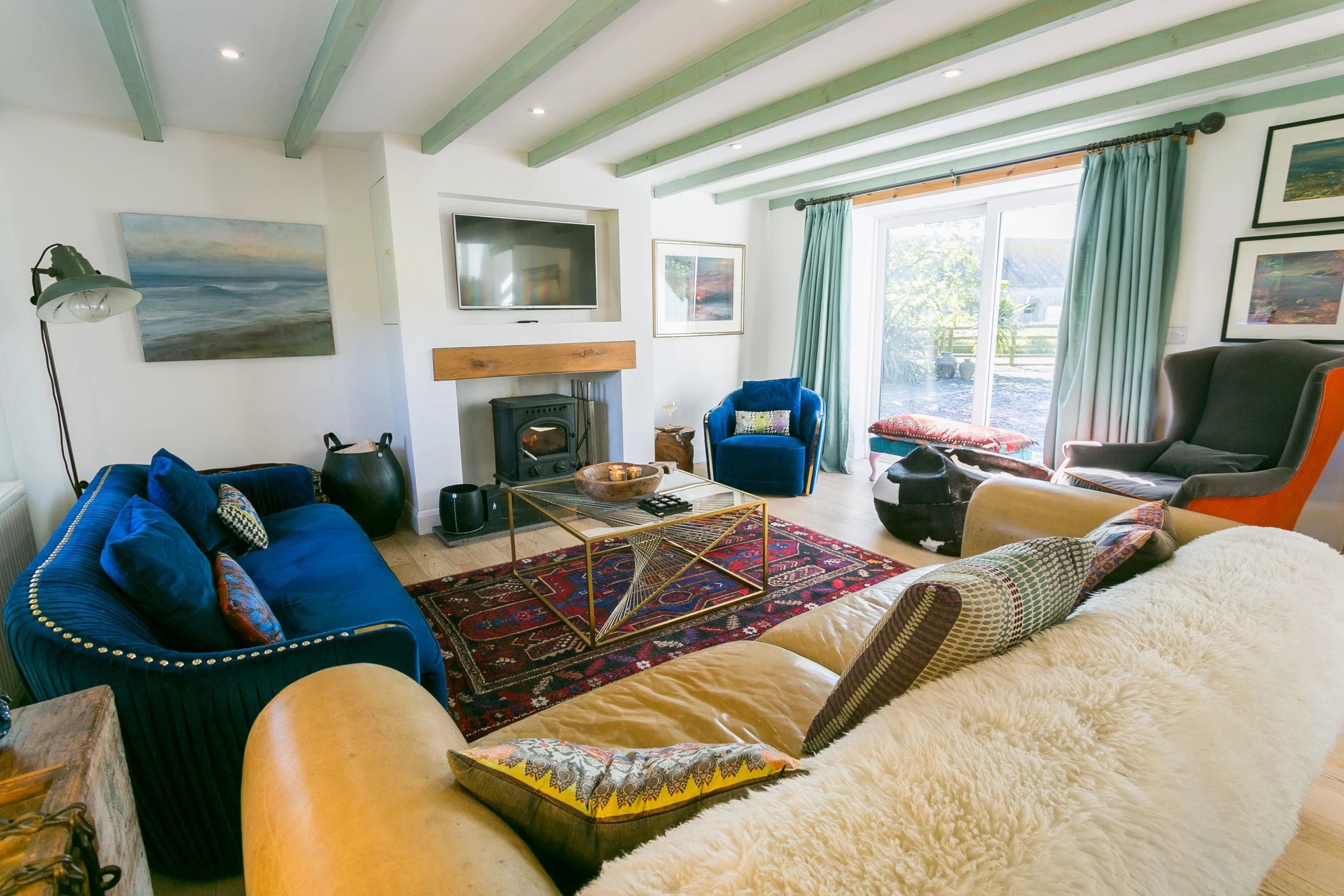 living room at Hen Felin showing wooden beamed ceiling, blue and yellow sofas, two arm chairs, a log burner and french doors. 