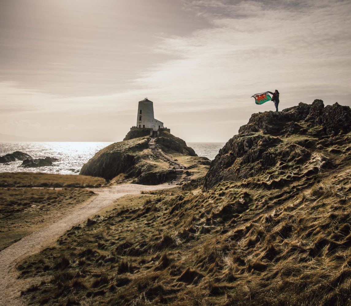 Newborough Warren walk. Shows two hills with a path between them and the Welsh flag is being held up by a woman on top of the hill on the right.  